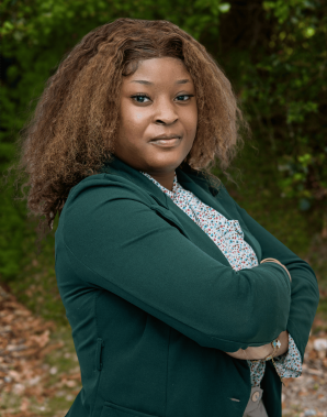 Meet the team: A woman with curly brown hair, wearing a green blazer and patterned shirt, stands outdoors with her arms crossed, looking confidently at the camera.