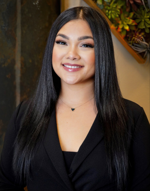 Meet the Team: A woman with long straight black hair, wearing a black blazer and necklace, smiles at the camera in an indoor setting with a plant wall in the background.