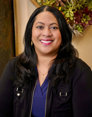 Meet the Team: A woman with long dark hair, wearing a black jacket over a blue top, smiles at the camera with a decorative plant wall in the background.